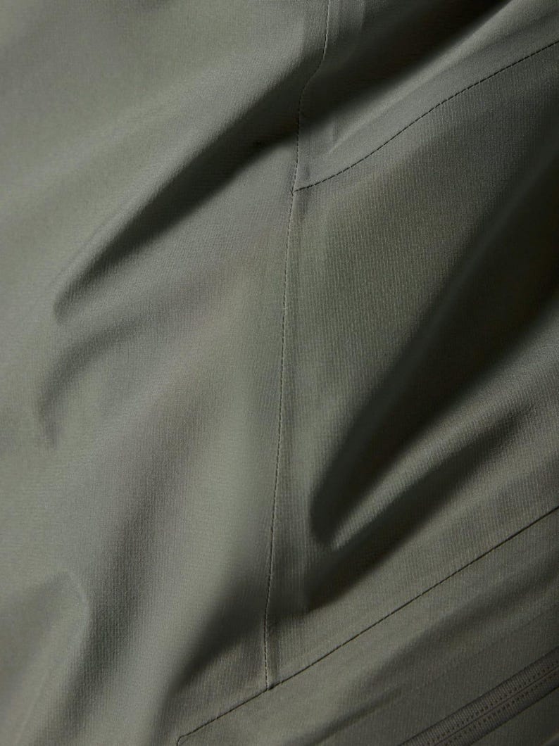 Puede incluir: Primer plano de tela verde oliva con textura y costuras visibles. La tela parece ser un material duradero, posiblemente impermeable, con sutiles variaciones de tono. La imagen sugiere una prenda de vestir o equipo para exteriores.