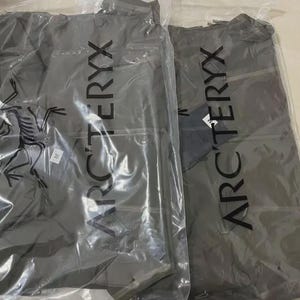 Może przedstawiać: Dwie szare kurtki w przezroczystym plastikowym opakowaniu. Kurtki mają pionowo nadrukowaną nazwę marki "ARC'TERYX". Logo marki, stylizowany skamieniały ptak, jest widoczne na jednej kurtce. Obie kurtki są nowe z metkami.