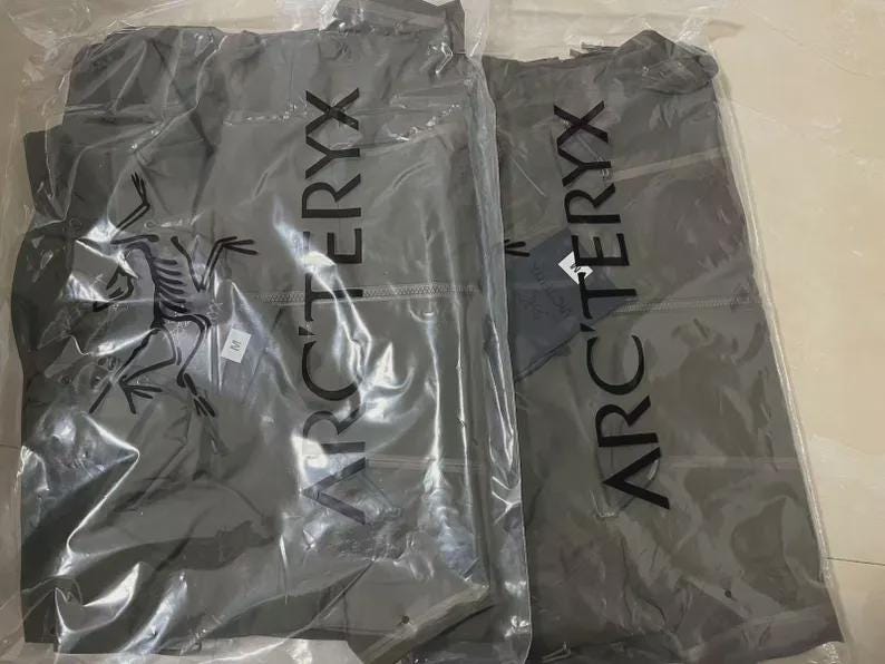 Może przedstawiać: Dwie szare kurtki w przezroczystym plastikowym opakowaniu. Kurtki mają pionowo nadrukowaną nazwę marki "ARC'TERYX". Logo marki, stylizowany skamieniały ptak, jest widoczne na jednej kurtce. Obie kurtki są nowe z metkami.