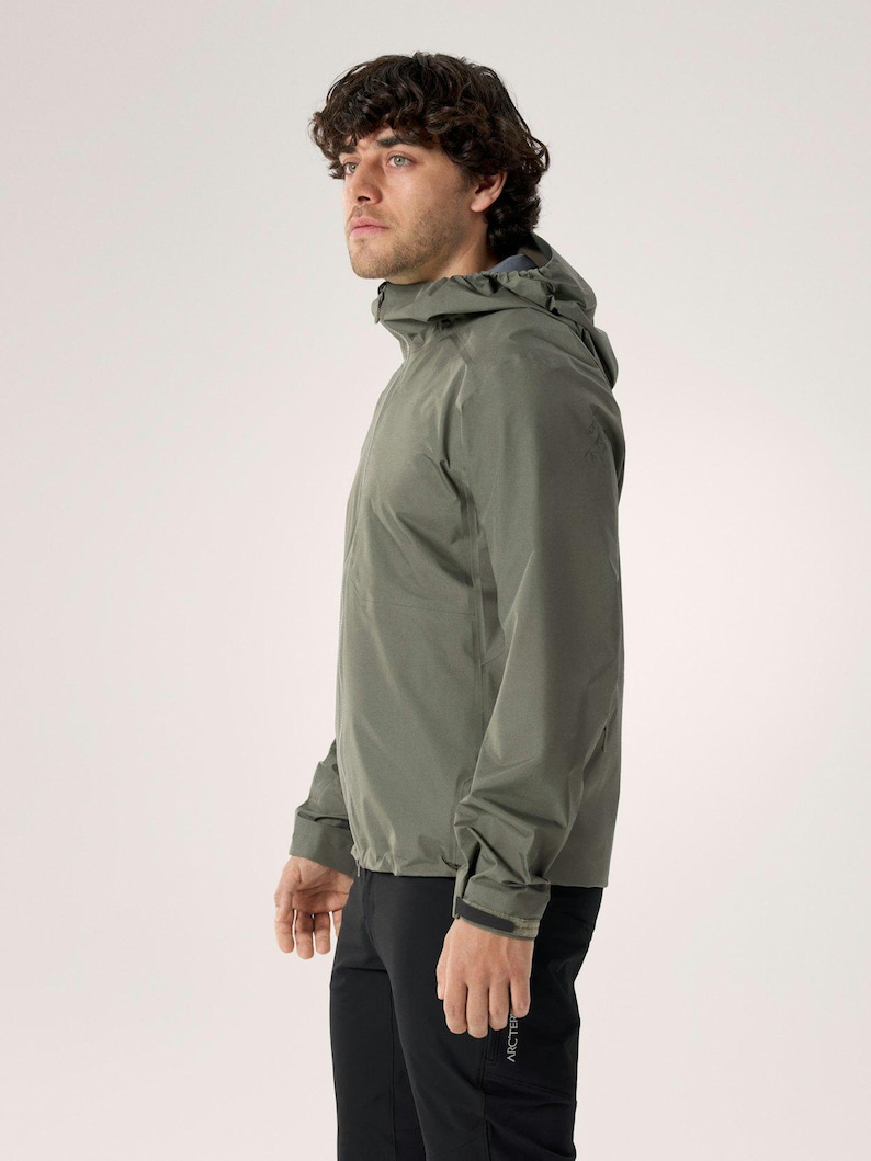 Puede incluir: Chaqueta con capucha verde oliva con mangas largas y cierre de cremallera. El modelo lleva pantalones negros. El modelo est&aacute; de perfil, mostrando el dise&ntilde;o de la chaqueta. Dise&ntilde;o minimalista.