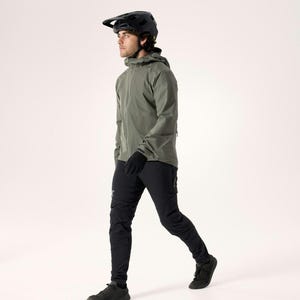 Puede incluir: Una persona con un casco negro, una chaqueta verde oliva, guantes negros y pantalones negros. La persona camina hacia adelante, con un fondo neutro. La chaqueta tiene una capucha y una cremallera. Los pantalones tienen un logotipo en la pierna izquierda. Los zapatos son negros.