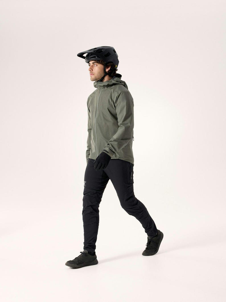 Puede incluir: Una persona con un casco negro, una chaqueta verde oliva, guantes negros y pantalones negros. La persona camina hacia adelante, con un fondo neutro. La chaqueta tiene una capucha y una cremallera. Los pantalones tienen un logotipo en la pierna izquierda. Los zapatos son negros.