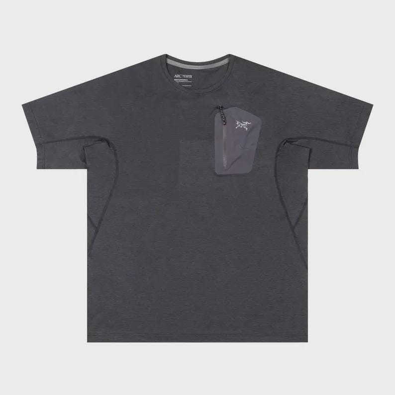 May include: Dark grey short-sleeve t-shirt with a small pocket on the chest. The pocket has a zipper and a white logo. The shirt has a crew neck and a subtle texture.