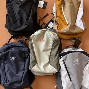 May include: Five backpacks in various colors: black, tan, navy blue, light green, and gray. Each backpack features a zippered main compartment, adjustable straps, and a logo on the front. The backpacks are made of a durable, water-resistant material.