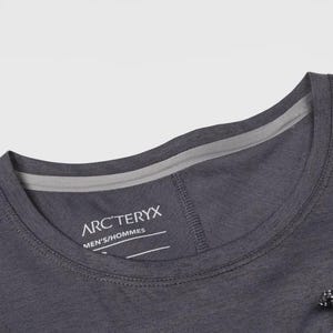May include: Close-up of a dark gray crew neck shirt. The shirt features the brand name "ARC'TERYX" and the text "MEN'S/HOMMES" printed on the inside collar. The shirt has a small pocket with a drawstring.