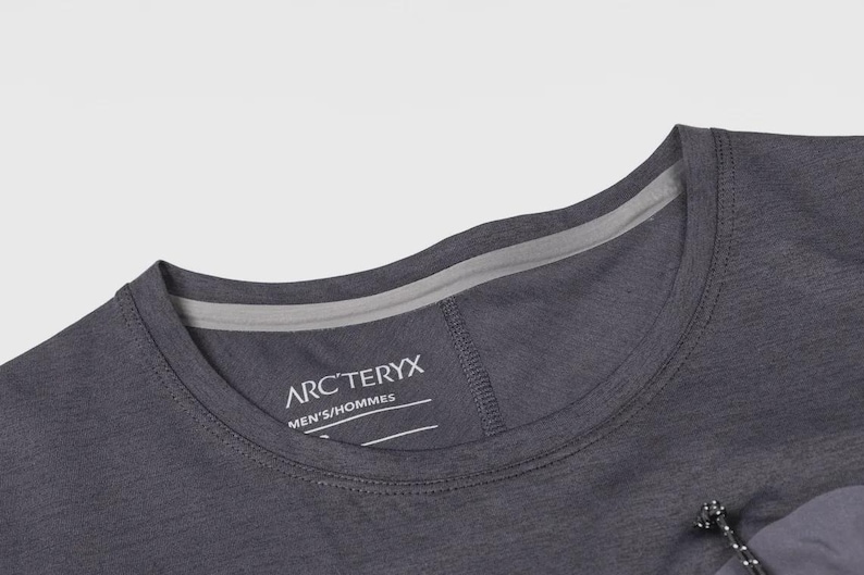 May include: Close-up of a dark gray crew neck shirt. The shirt features the brand name "ARC'TERYX" and the text "MEN'S/HOMMES" printed on the inside collar. The shirt has a small pocket with a drawstring.