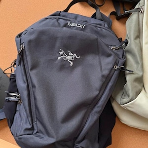 May include: A dark gray backpack with the brand name "ARCTERYX" and a white logo. The backpack has a top handle, two zippered compartments, and adjustable straps. Another backpack is partially visible in the background.