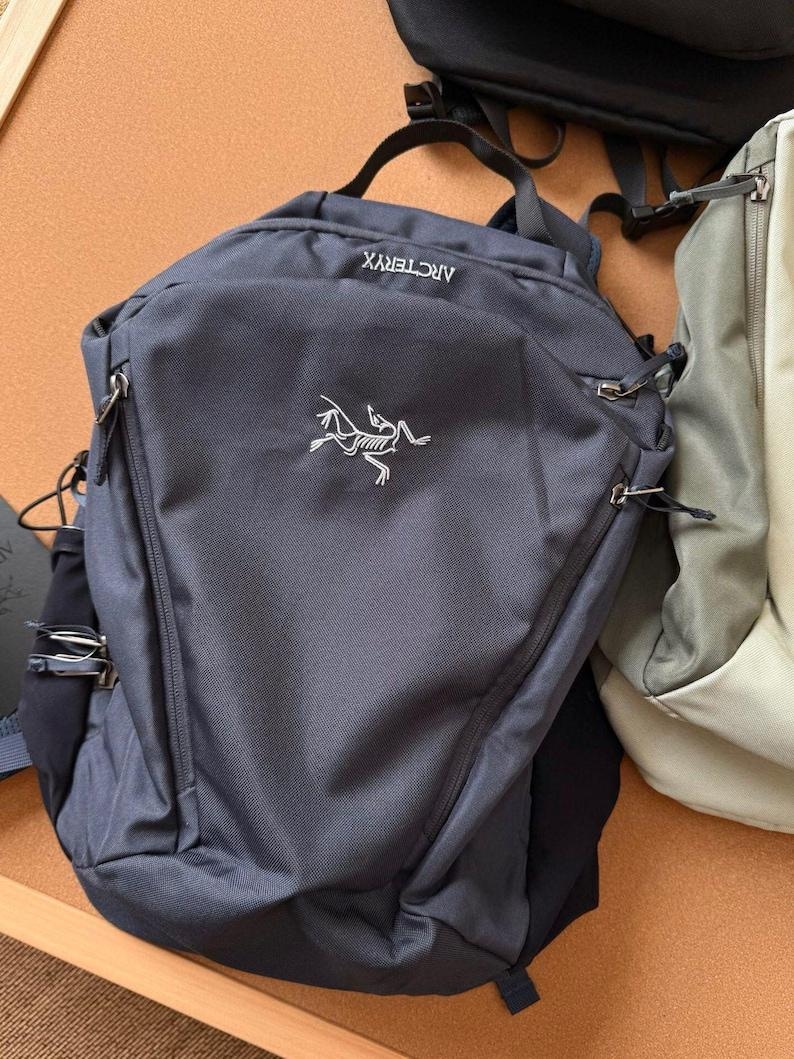 May include: A dark gray backpack with the brand name "ARCTERYX" and a white logo. The backpack has a top handle, two zippered compartments, and adjustable straps. Another backpack is partially visible in the background.