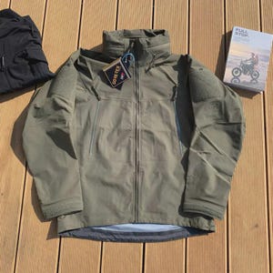 Może przedstawiać: Zielona kurtka w kolorze oliwkowym z wysokim kołnierzem, zapięciem na zamek i wieloma kieszeniami. Kurtka ma dołączoną metkę Gore-Tex. Po lewej stronie widoczny jest czarny element garderoby. Po prawej książka z tytułem "FULL STOP".
