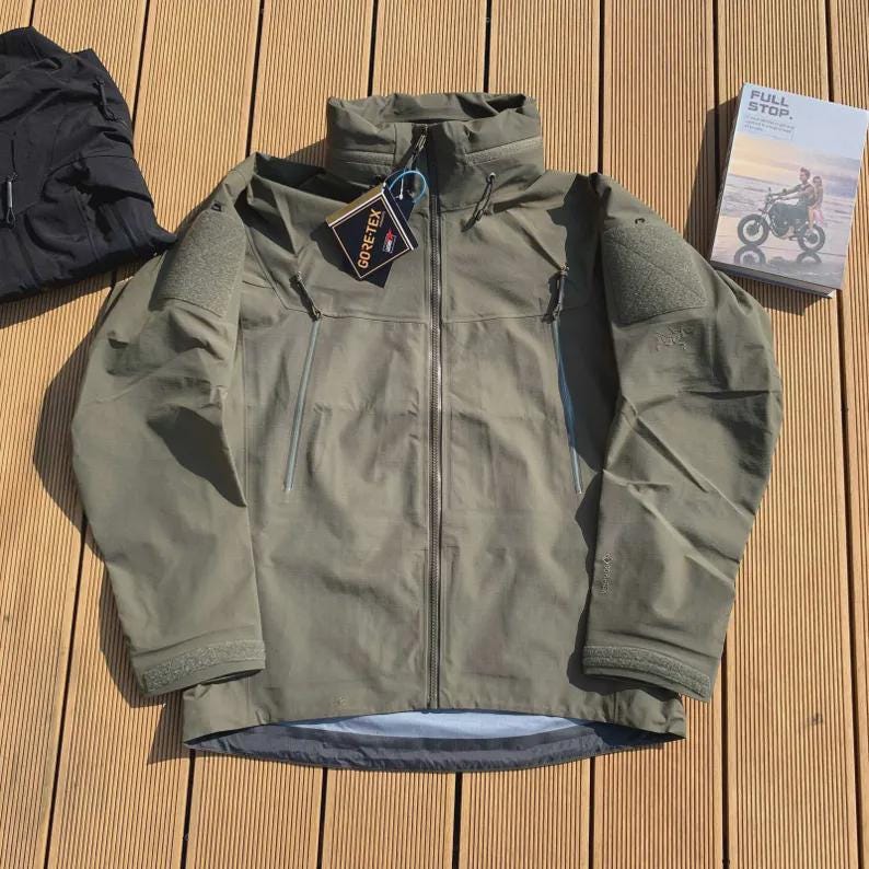 Może przedstawiać: Zielona kurtka w kolorze oliwkowym z wysokim kołnierzem, zapięciem na zamek i wieloma kieszeniami. Kurtka ma dołączoną metkę Gore-Tex. Po lewej stronie widoczny jest czarny element garderoby. Po prawej książka z tytułem "FULL STOP".