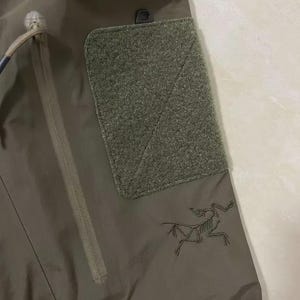 Może przedstawiać: Zbliżenie na oliwkowozielone spodnie outdoorowe. Zawierają kieszeń na zamek błyskawiczny, naszywkę na rzep i czarne haftowane logo stylizowanego szkieletu zwierzęcia. Spodnie wykonane są z wytrzymałego, wodoodpornego materiału.