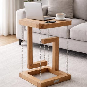 Puede incluir: Una mesa auxiliar de madera con un diseño flotante único. La mesa es de madera clara y está suspendida por cadenas plateadas. Una computadora portátil, un teléfono inteligente y una taza de café están sobre la mesa. La mesa mide aproximadamente 61 cm de largo, 41 cm de ancho y 51 cm de alto.