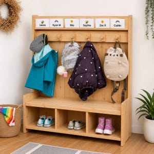 May include: A light wood entryway storage bench with six hooks, a top shelf with name labels Lewis, Ryan M, Mia, Sophie, Zac R, and Caitlin, and three bottom cubbies. Items hanging include a teal jacket, a glittery backpack, a dark coat, and a cat bag.