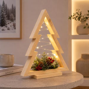 May include: A triangular wooden frame shaped like a Christmas tree, featuring a light-coloured natural wood finish. The centre is illuminated by thin, glowing string lights and decorated at the base with artificial pine needles, pinecones, and red berries.