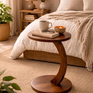 May include: A dark brown, curved side table with a round top and base. A mug, book, and glasses rest on the table. A lit candle in a small bowl sits nearby. The table is next to a bed with a white comforter.