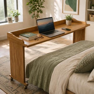 May include: A wooden laptop desk with a raised edge, positioned over a bed. The desk holds a laptop, phone, books, and glasses. The bed has a green blanket and pillows. The desk is on wheels.