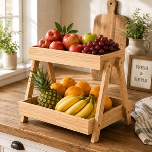 May include: A two-tiered wooden fruit stand filled with fresh produce. The top level holds red apples, green grapes, and a green apple. The bottom level contains oranges, bananas, and a pineapple. The stand is made of light-colored wood.