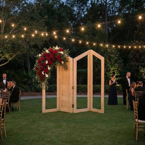 May include: A wooden folding screen with an arch of red roses and white flowers, set up outdoors for a wedding ceremony. The screen is positioned on a green lawn, with guests and string lights in the background.