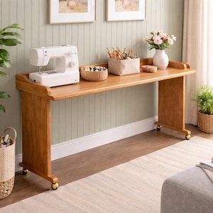 May include: A wooden sewing table with a white sewing machine, a basket of thread, and a beige fabric container filled with sewing supplies. The table has a long, flat surface and is supported by wooden legs with gold-colored wheels.