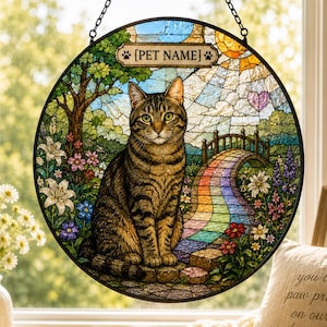 Op de afbeelding: Een ronde glas-in-lood zonnevanger met een bruine cyperse kat die in een tuin zit met een regenboogbrug op de achtergrond. Een rechthoekig plaatje bovenaan bevat de tekst [PET NAME], geflankeerd door twee kleine pootafdruk-pictogrammen.