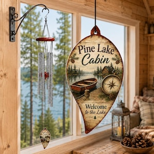May include: A wind chime with silver metal tubes and a small teardrop-shaped sign hanging from a black metal wall bracket. Beside it hangs a large teardrop-shaped wooden sign featuring a lake scene, a boat, a compass, and the text Pine Lake Cabin.