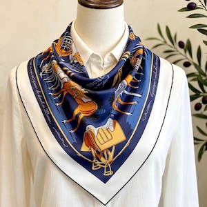 May include: A square silk scarf featuring a deep blue background with orange and white illustrations of horse saddles, stirrups, and equestrian gear. The scarf has a wide white border with thin black piping and cursive text along the edges.