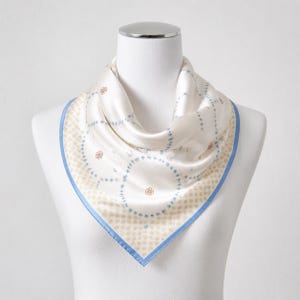 May include: A square silk scarf in cream featuring a light blue border and a repeating pattern of small tan dots. The center displays interconnected light blue circular chains and small brown floral motifs printed on the smooth fabric surface.