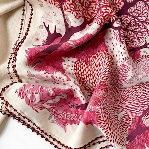 May include: A cream-colored fabric scarf featuring a detailed print of trees, foliage, and birds in shades of red, pink, and burgundy. The edges are finished with a decorative dark red crocheted trim, adding texture to the patterned textile design.