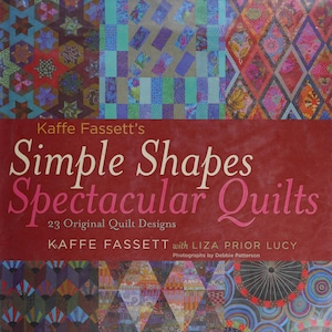 May include: A book titled Kaffe Fassett's Simple Shapes Spectacular Quilts with 23 Original Quilt Designs by Kaffe Fassett with Liza Prior Lucy. The cover features a deep red center with white and pink text, surrounded by colorful quilt patterns.