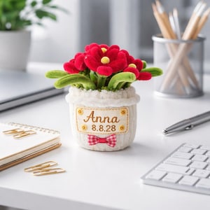 May include: A plush, white flower pot with red flowers and green leaves. The pot has a label embroidered with the name "Anna" and the date "8.8.28", and a red and white gingham bow. The pot is on a white desk.