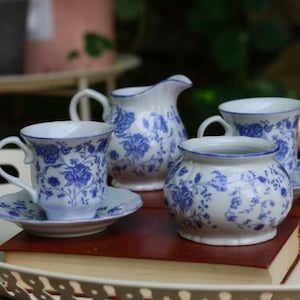 Pode incluir: Um conjunto de chá em porcelana branca com um padrão floral azul. O conjunto inclui uma leiteira com pega e bico, um açucareiro e duas chávenas de chá com pires a condizer. Cada peça apresenta um rebordo azul delicado e motivos florais.