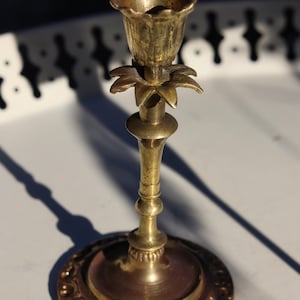 May include: A brass candlestick holder featuring a flower-shaped bobeche and a tulip-shaped candle cup. The stem has a slender, tapered design with a circular base decorated with an embossed scrollwork pattern along the outer edge.