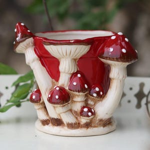 May include: A ceramic planter featuring a sculpted design of multiple red mushrooms with white spots and cream-coloured stalks. The exterior is glossy, showcasing a cluster of fungi of varying sizes wrapping around the cylindrical vessel body.