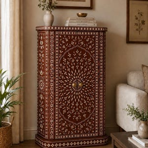 May include: A tall, rectangular wooden cabinet featuring intricate white floral and geometric inlay patterns across its dark brown surface. The cabinet has two doors with small, round gold-toned knobs and rounded corners, showcasing detailed craftsmanship.
