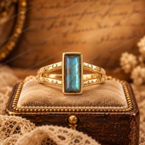 May include: A gold ring featuring a rectangular, faceted labradorite gemstone with blue iridescence. The band is split and decorated with engraved sun and moon motifs along the sides, framing the central stone in a polished gold setting.