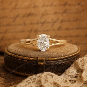 May include: A gold engagement ring with an oval-cut diamond. The ring has a split shank design, with the band splitting on either side of the diamond. The ring is set on a brown velvet jewellery box, with a lace cloth in the background.