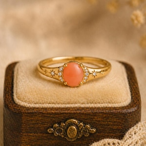 May include: A gold ring featuring a central oval-shaped peach-colored stone, flanked by small clear stones set within star-shaped engravings on the band. The ring has a polished finish and a delicate design with symmetrical star accents on both sides.