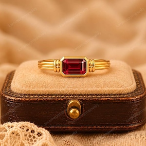 Puede incluir: Un anillo de oro con una gema rectangular de color rojo intenso. El anillo tiene una banda de oro con detalles acanalados a los lados de la gema. El anillo se exhibe en un joyero marrón con un cierre dorado.
