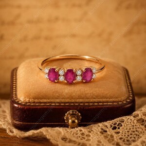 May include: A gold ring featuring three oval-cut, vibrant red gemstones, flanked by smaller, clear stones. The ring is displayed on a velvet cushion inside a vintage jewelry box, showcasing its elegant design and craftsmanship. The ring is a piece of fine jewelry.