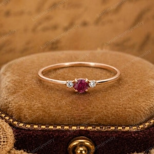 Könnte beinhalten: Ein zarter roségoldener Ring mit einem zentralen runden Rubin-Edelstein, flankiert von zwei kleineren, klaren Edelsteinen. Der dünne Ringband präsentiert das leuchtende Rot des Rubins. Ein klassisches Schmuckstück.