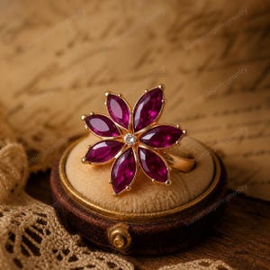 May include: A gold ring featuring a flower design with six deep red marquise-cut gemstones as petals. A small, round diamond is set in the center. The ring is displayed on a velvet cushion inside a small, round jewelry box.