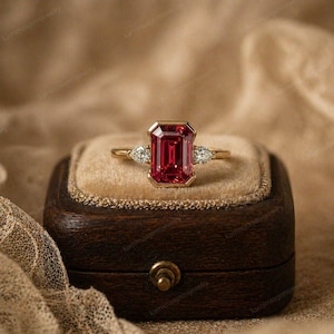 May include: A gold ring featuring a large, rectangular, red gemstone as the centerpiece, flanked by two smaller, pear-shaped, clear gemstones. The ring is set on a brown velvet surface inside a wooden jewelry box.