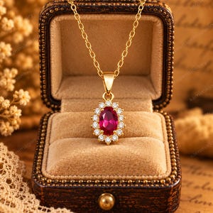 May include: A gold chain necklace featuring an oval-cut vibrant red gemstone pendant surrounded by a halo of small, clear, round-cut white stones. The pendant is displayed inside a brown, velvet-lined jewelry box with a hinged lid.