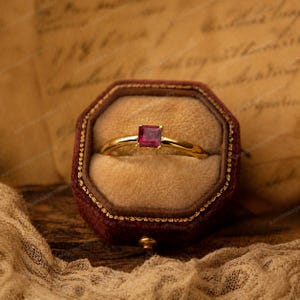 May include: A gold ring with a square, deep red gemstone is displayed inside a burgundy velvet ring box. The ring has a simple, elegant design, with the gemstone set in a minimalist setting. The box is octagonal.