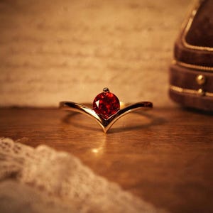 May include: A gold-colored ring with a V-shaped band and a large, round, red gemstone. The gemstone has a small, round accent on top. The ring is set on a wooden surface.