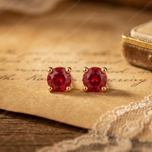 May include: A pair of round-cut red gemstone stud earrings set in gold metal. Each earring features a faceted circular red stone secured by four small gold prongs, creating a classic stud design with a vibrant, deep red color and metallic gold finish.