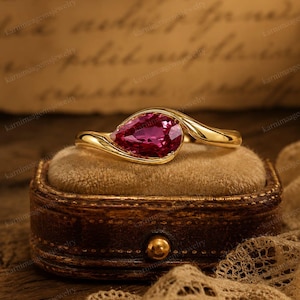 May include: A gold ring featuring a pear-shaped, deep pink gemstone set in a bypass-style band. The ring is positioned on a velvet-lined, dark brown, oval-shaped container. The metal band has a smooth, polished finish that curves around the stone.