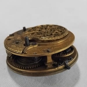 May include: Close-up of a vintage watch movement, showcasing intricate gears and components. The brass-colored metal is detailed with decorative patterns. The watch mechanism is partially disassembled, revealing the internal workings of the timepiece.