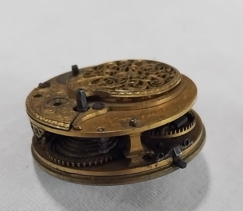 May include: Close-up of a vintage watch movement, showcasing intricate gears and components. The brass-colored metal is detailed with decorative patterns. The watch mechanism is partially disassembled, revealing the internal workings of the timepiece.