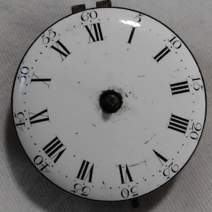May include: A white clock face with black Roman numerals and minute markers. The clock face has a black outer ring and a central black mechanism. The numerals are arranged around the perimeter of the face.